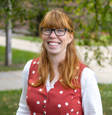 Dr. Bridget Moricz smiles at the camera. She has long red hair, dark glasses, and is wearing a red and white polka dot vest over a white shirt. 