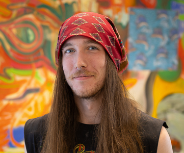 Bryce Waller smiles at the camera. He is standing in front of an extremely colorful painting.