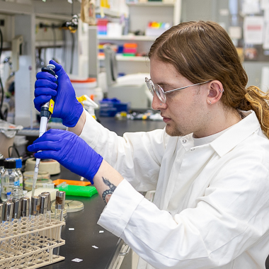 Alex Wessel works at his bench. He's got long brown hair, glasses, and is wearing a lab coat and blue gloves. He's pipetting. 