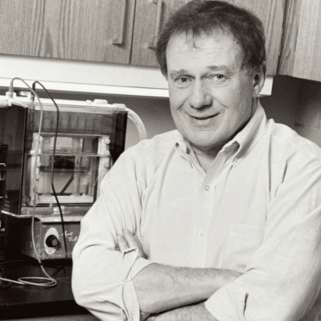 This image shows Dr. Bob Brubaker sitting in front of cabinets and some type of equipment. He is wearing a button downs hirt and is smiling. 