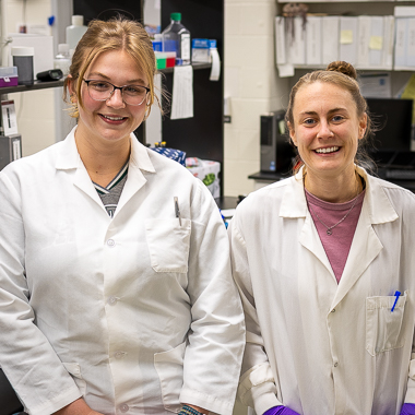 Kyleen Hall and Danielle Gregory stand in their lab. They are smiling at the camera and both are wearing lab coats. 