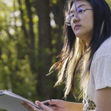 Faith Nhkum draws outside. The sun is catching her hair, and there are trees shown in bokeh in the background. 