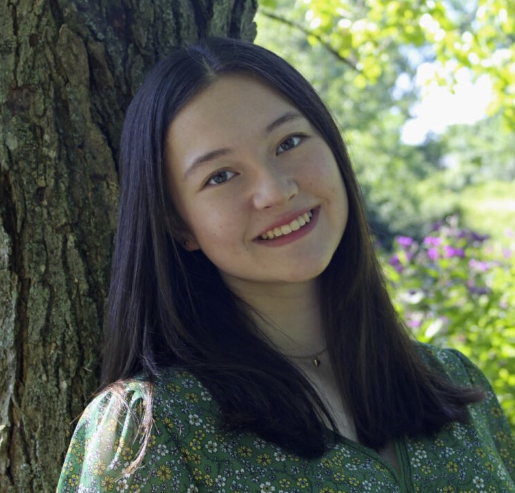Girl with long black hair smiles at the camera. She is leaning against a tree. 